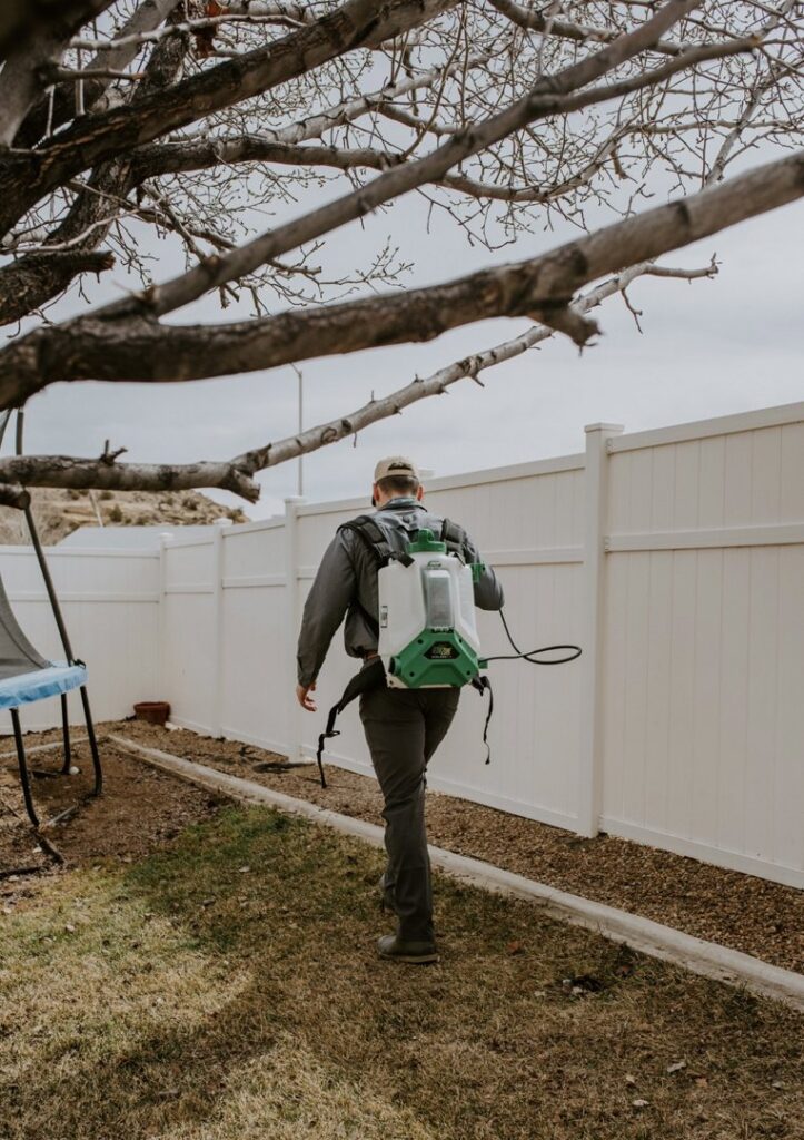 A Box Pest Control technician walking through a backyard with a sprayer, performing pest control in Farmington, NM.