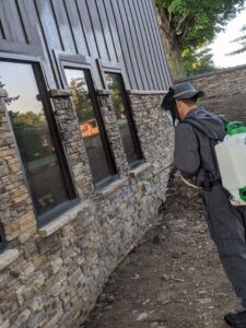 A technician in protective gear sprays the exterior of a building, performing pest control for Optimum Pest Pros, LLC. in Canandaigua, NY.