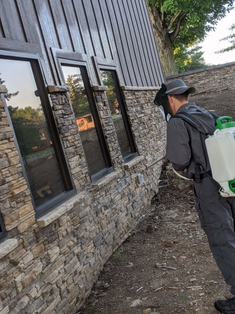 A technician in protective gear sprays the exterior of a building, performing pest control for Optimum Pest Pros, LLC. in Canandaigua, NY.