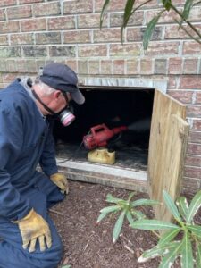 A Canady & Son Exterminating technician in protective gear spraying into a crawl space opening for pest treatment in Wilmington, NC.