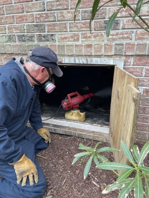 A Canady & Son Exterminating technician in protective gear spraying into a crawl space opening for pest treatment in Wilmington, NC.