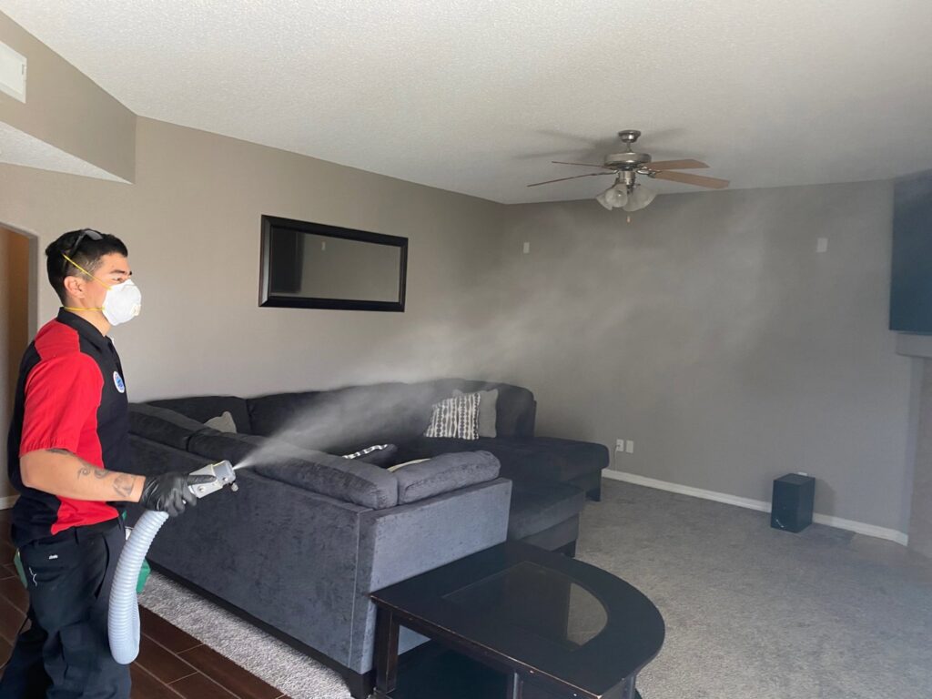 A technician in a mask spraying deodorizer in a living room as part of a service by Steamway Carpet Cleaning in El Paso, TX