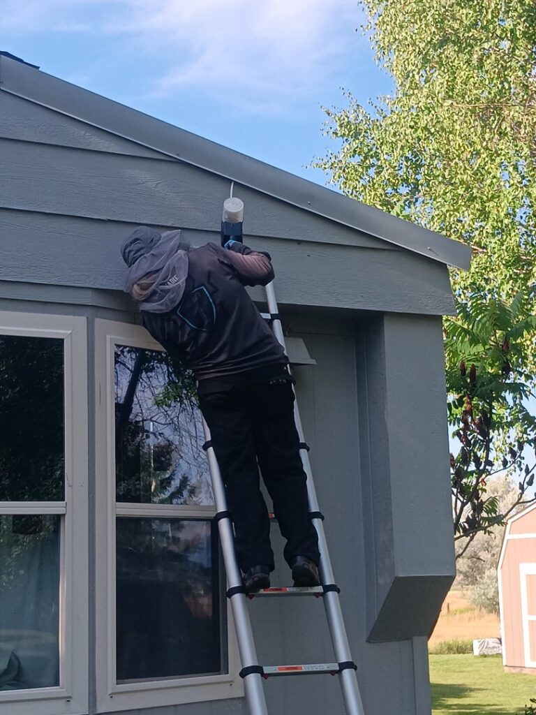 A Pest One technician on a ladder spraying the eaves of a house for pest control in Bozeman, MT.