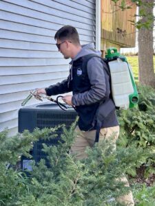 A pest control technician using a backpack sprayer to treat the exterior of a house near an AC unit for Modern Solutions Pest Control in Rockford, IL.