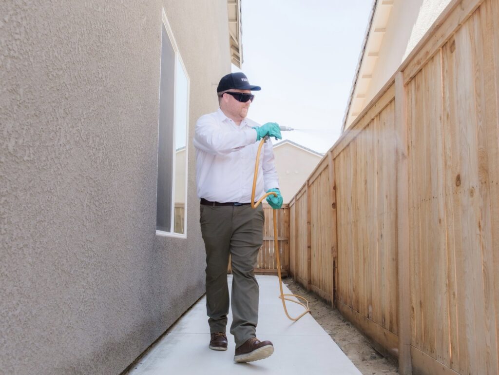 A Thrive Pest Pros technician spraying pest control treatment along a fence line next to a home in Clovis, CA.