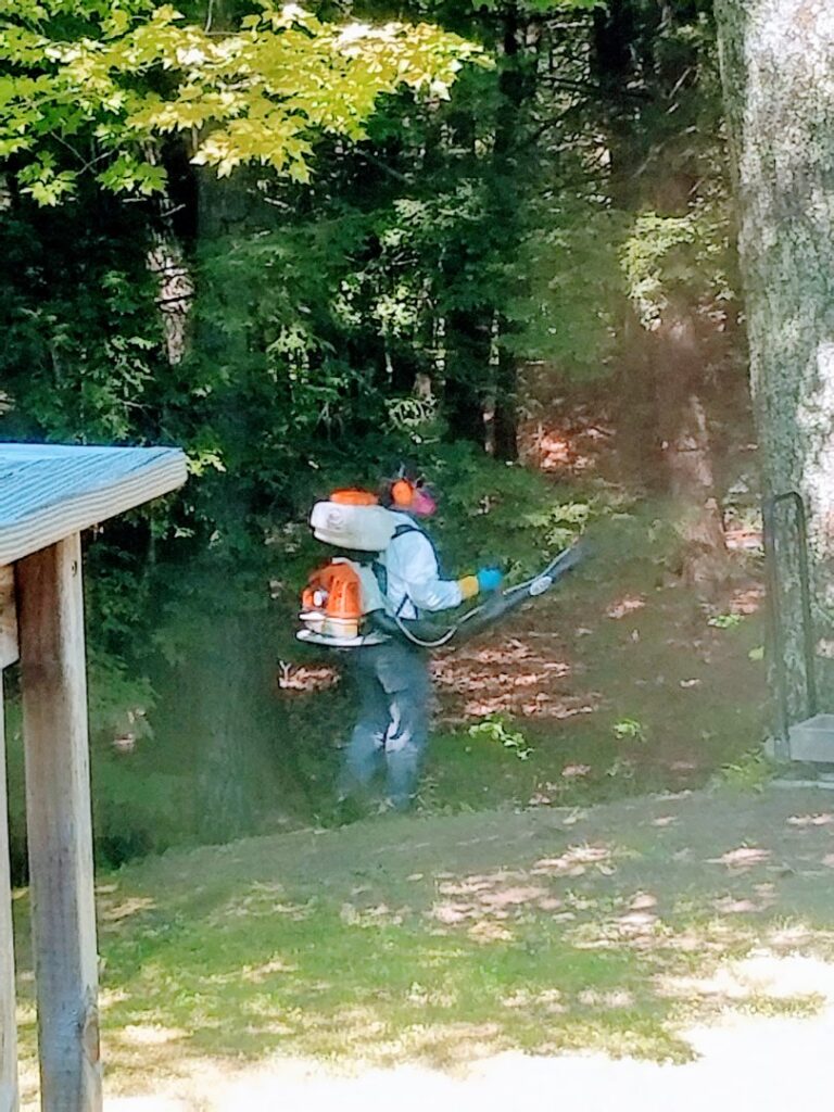 A pest control technician in protective gear using a backpack sprayer in a wooded area for Pine State Pest Solutions in Auburn, ME.