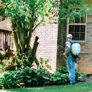 A technician from Liberty Pest Services LLC spraying the exterior of a brick house and bushes with a backpack sprayer in Mayflower, AR.
