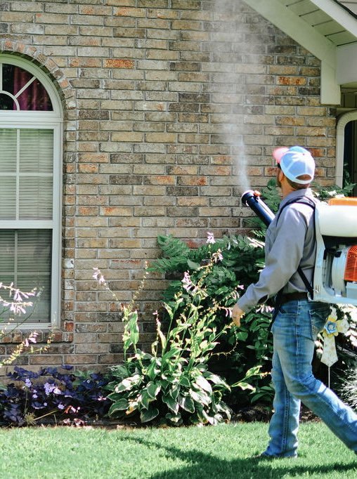 A technician from Liberty Pest Services LLC spraying the exterior of a brick house and surrounding bushes in Mayflower, AR.