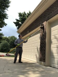 A Bayer Pest Control technician spraying the exterior of a house near the garage door with a long pole for pest treatment in Dayton, OH.