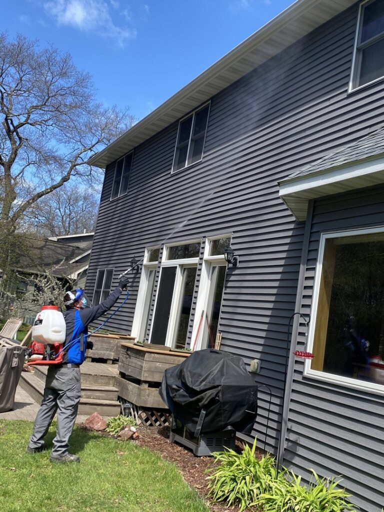 A pest control technician applying treatment to the exterior of a house with a backpack sprayer for 1st Choice Pest Solutions in Eau Claire, WI.