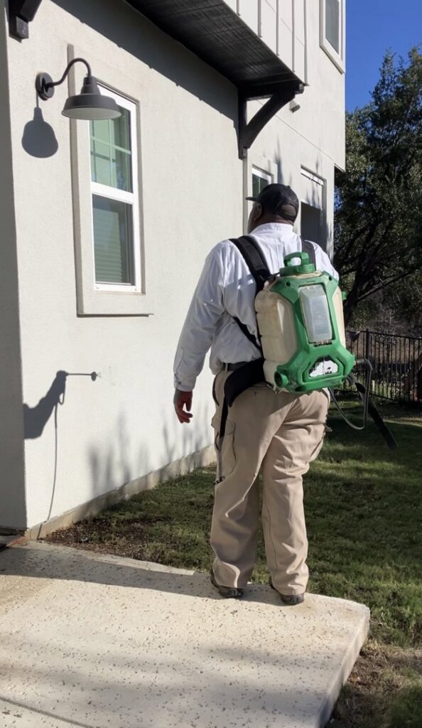 A pest control technician from A-TEX PEST MANAGEMENT INC. in Austin, TX, applying treatment to the exterior of a house with a backpack sprayer