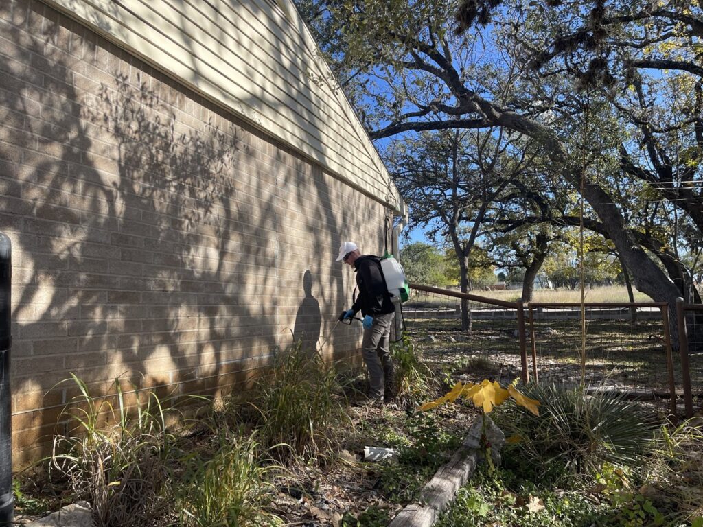 A Blessed Pest Control technician applying an exterior pest treatment to the foundation of a brick house in San Antonio, TX