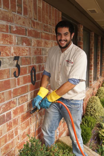 A smiling technician spraying the foundation of a brick house for pest control by Reliable Termite of OKC in Oklahoma City, OK.