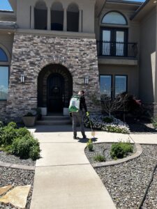 A Box Pest Control technician spraying the perimeter of a house and garden beds for pest control in Farmington, NM.