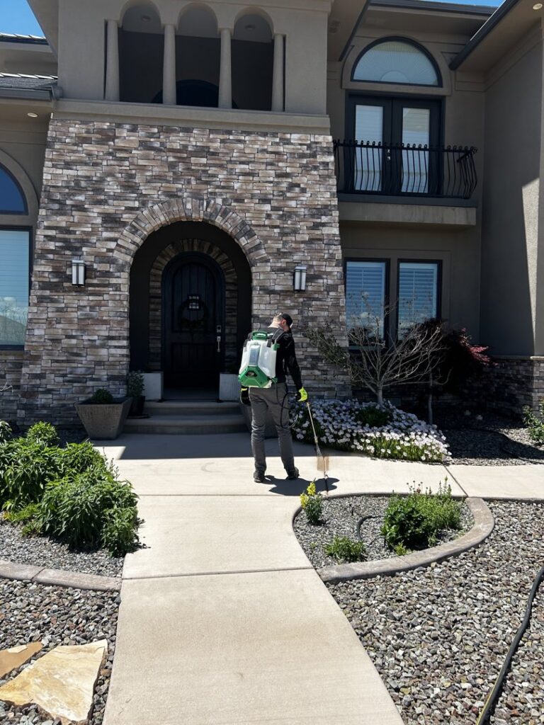 A Box Pest Control technician spraying the perimeter of a house and garden beds for pest control in Farmington, NM.