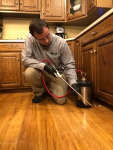 A Bayer Pest Control technician kneeling and applying pest treatment with a handheld sprayer along the kitchen floor in Dayton, OH.