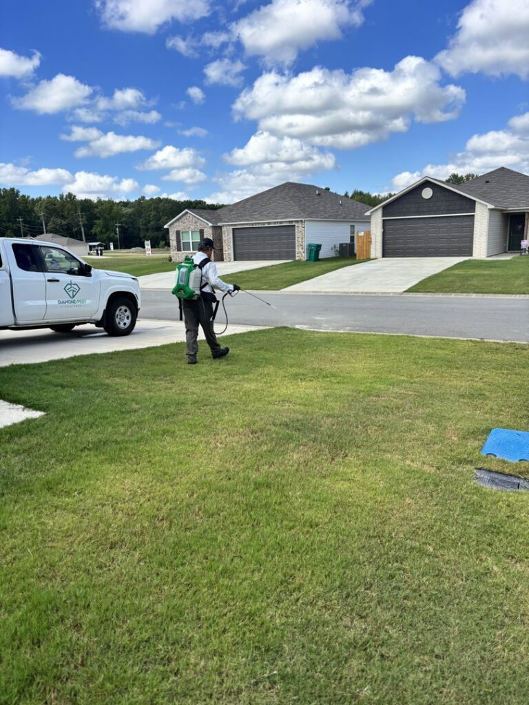 A Diamond Pest Solutions technician in Conway, AR, spraying a residential lawn as part of a pest control service, with a company truck nearby.