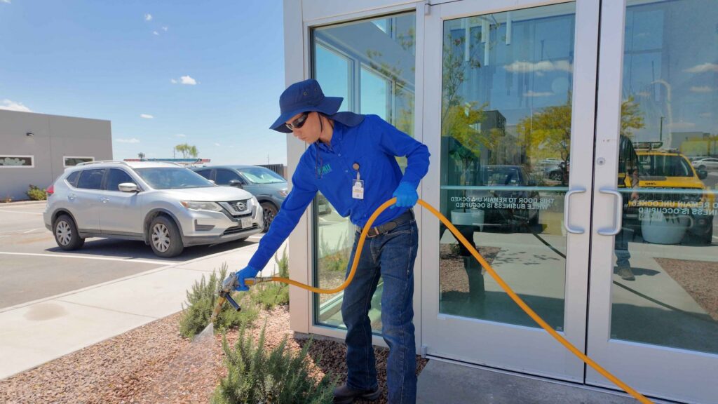 A technician from El Valle Pest Control spraying a pest treatment around the foundation of a commercial building in El Paso, TX