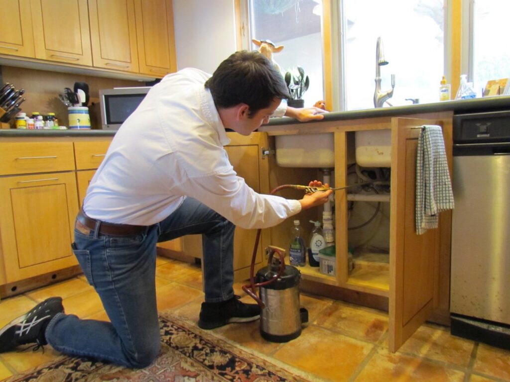 A pest control technician spraying under a kitchen sink during a service call for Roberts Termite & Pest Control in Austin, TX