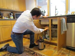 A pest control technician spraying under a kitchen sink during a service call for Roberts Termite & Pest Control in Austin, TX