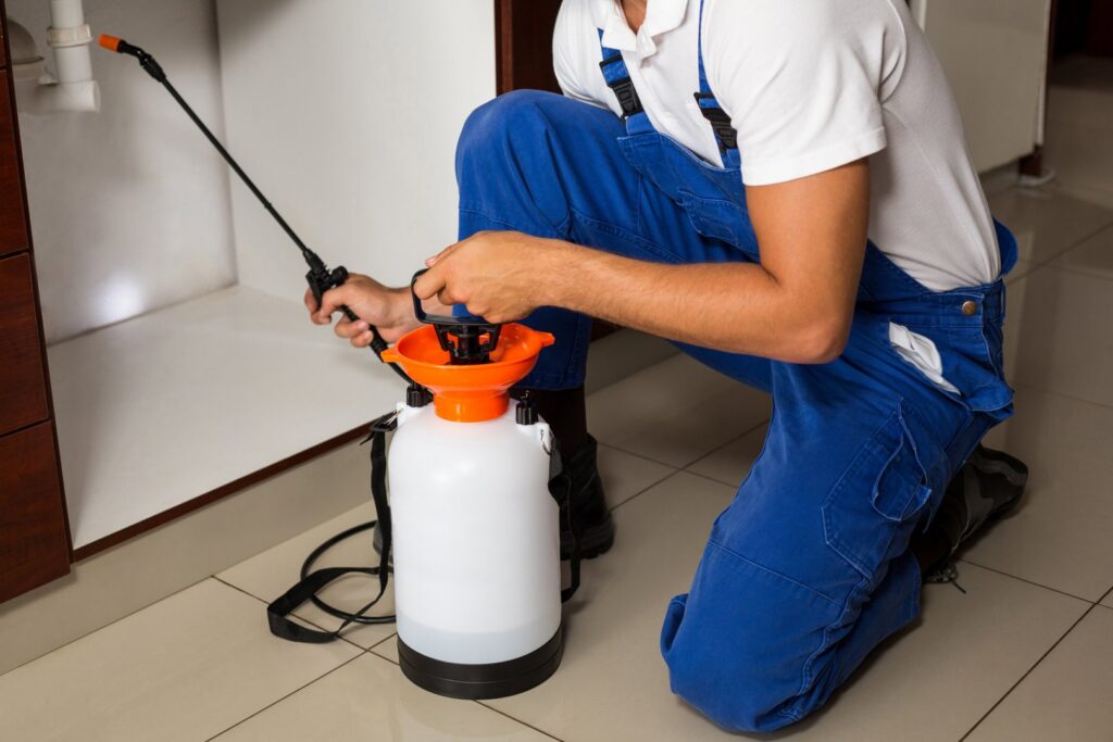 A pest control technician from Fusion Pest Control in Ammon, ID, kneeling and spraying under a kitchen sink to eliminate pests.