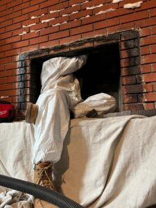 A Chimney Wizards LLC technician in a protective suit sweeping a fireplace, with a vacuum hose for cleanup in Orofino, ID.