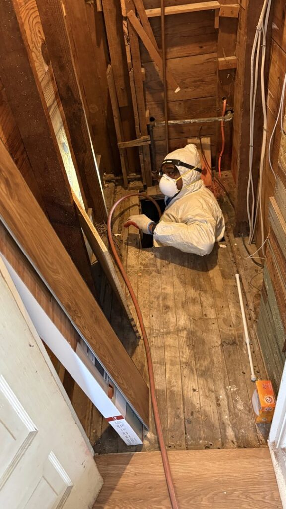 A D' Bug Man Pest Control technician in a protective suit treating a crawl space under a house in Arlington, TX.
