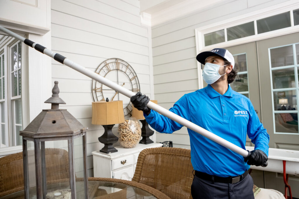 A Pest Authority Des Moines technician treating an exterior window or eave with a long pole tool in Des Moines, IA.