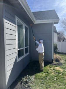 A Hemi Pest Control technician treating the exterior of a house with a long pole tool in Meridian, ID.