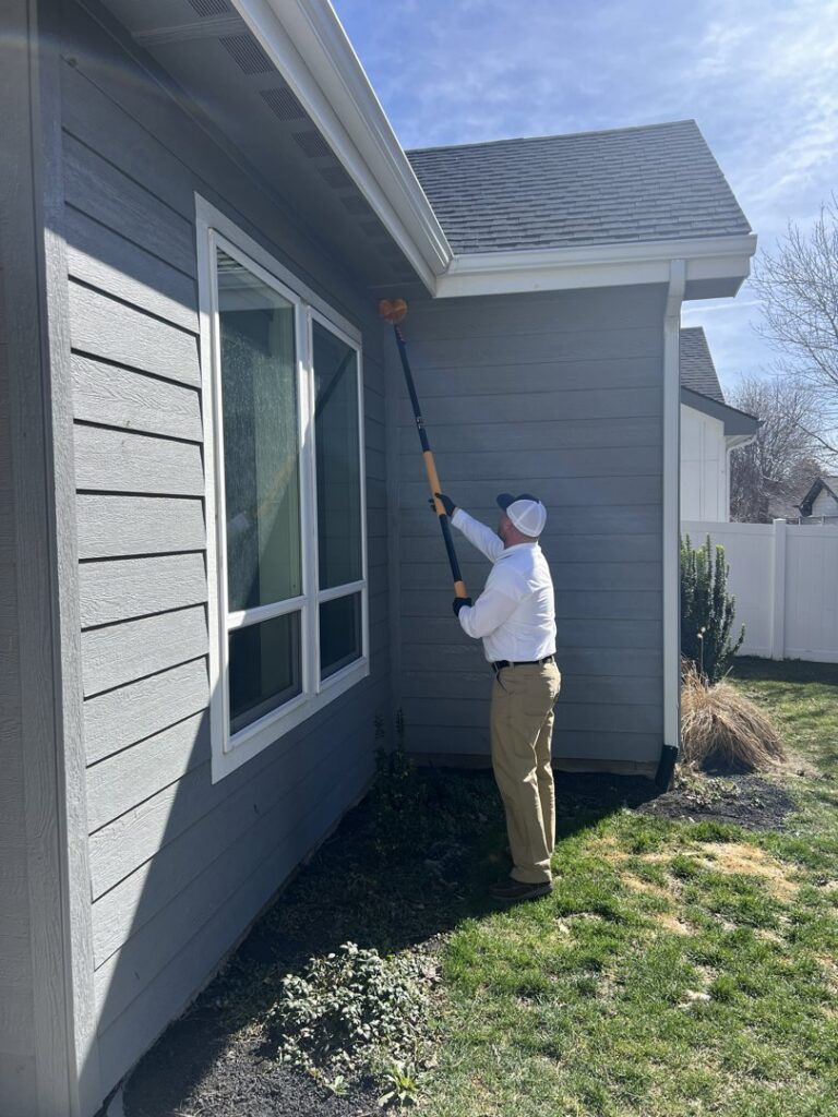 A Hemi Pest Control technician treating the exterior of a house with a long pole tool in Meridian, ID.