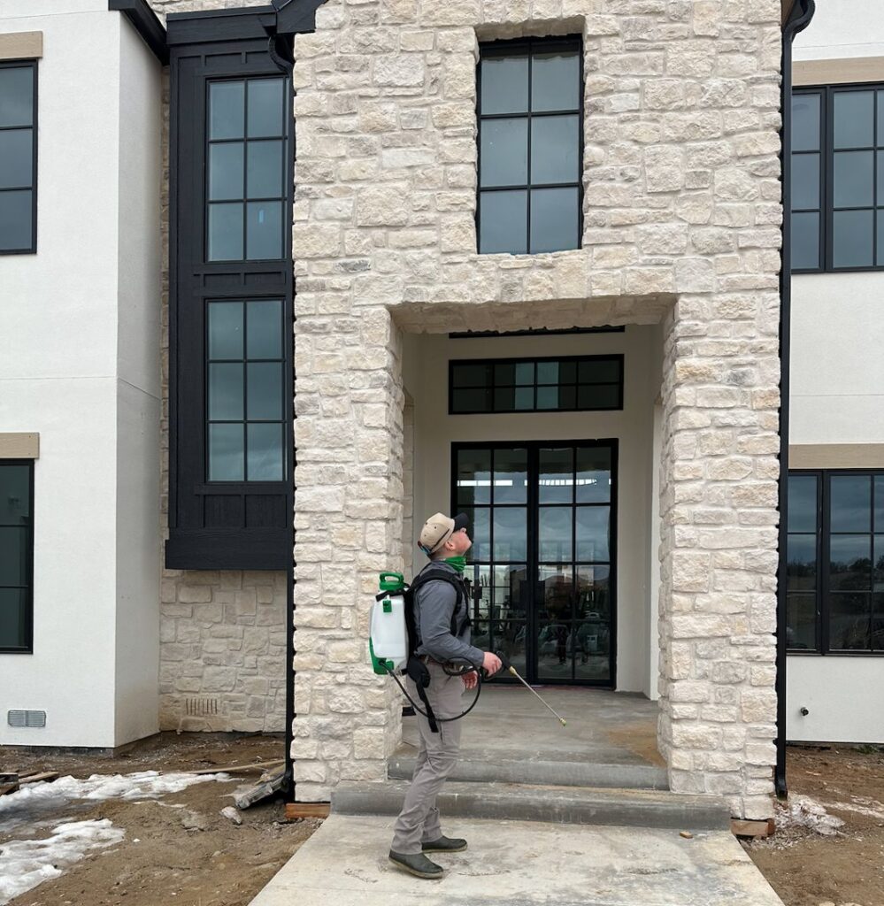 A Box Pest Control technician treating the foundation and entrance of a large home for pests in Farmington, NM.