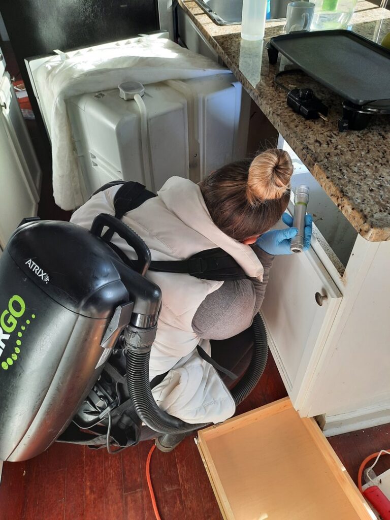 A technician using a backpack vacuum to clean under a kitchen cabinet, part of pest control cleanup by Direct Pest Solutions Of Georgia, LLC in McDonough, GA.