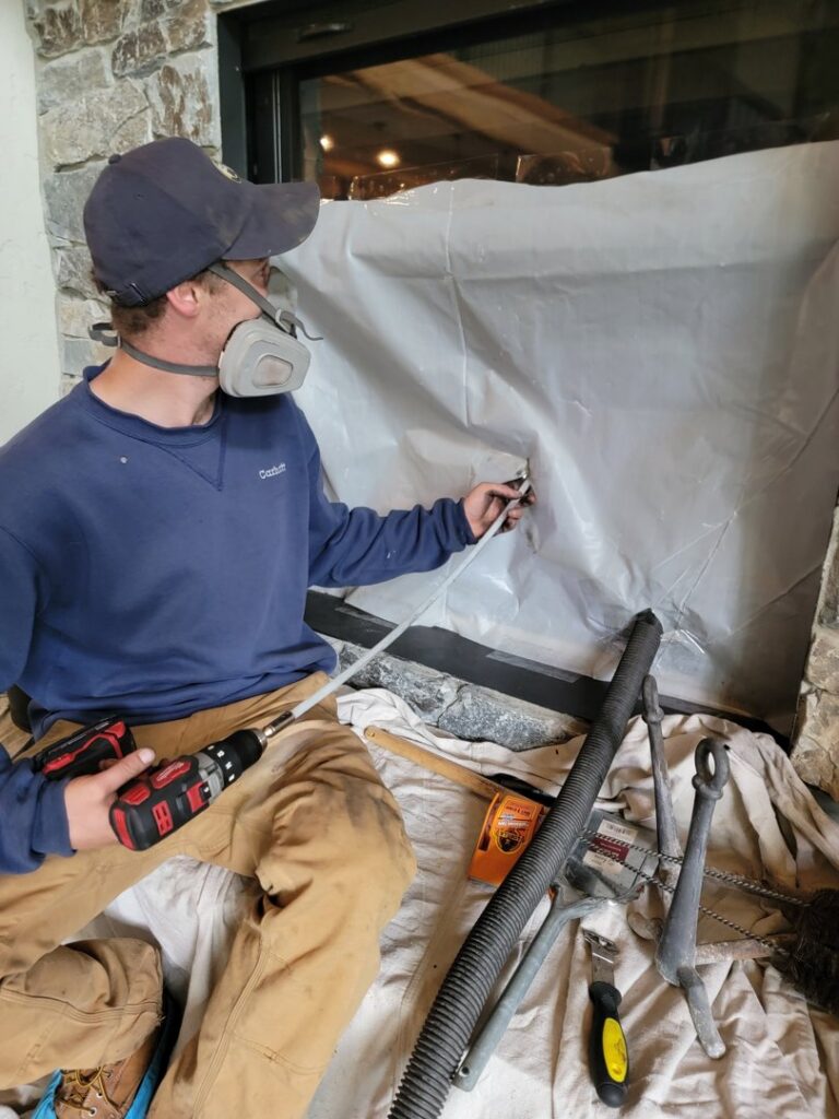 A Chimney Wizards LLC technician wearing a respirator mask while sweeping a fireplace, with plastic sheeting for dust control in Orofino, ID.