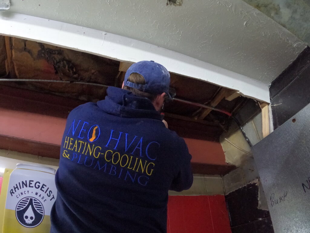 A technician from North East Ohio Master Plumbing, Heating, and Cooling Contractors working on pipes and ducts in a ceiling in Cleveland, OH.