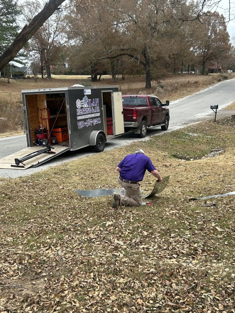A Milan Heating & Air technician working with sheet metal for an HVAC project next to a service trailer in Milan, TN.
