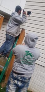 Two Clean Air Solutions technicians cleaning an exterior vent on a house, one on a ladder, in Gaithersburg, MD.