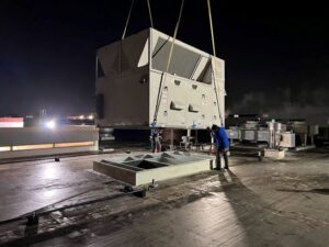 Two technicians from Deljo Heating, Cooling & Plumbing installing a large commercial HVAC unit on a rooftop at night in Chicago, IL.