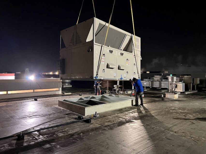 Two technicians from Deljo Heating, Cooling & Plumbing installing a large commercial HVAC unit on a rooftop at night in Chicago, IL.