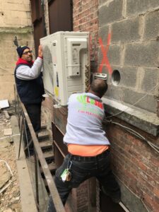 Two technicians from L&SON Heat AC Tech installing an outdoor mini-split air conditioning unit on a brick wall in Yonkers, NY.