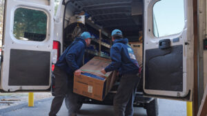 Two Blue Bear Plumbing, Heating, & Air technicians loading an HVAC unit into a service van in Norwell, MA.