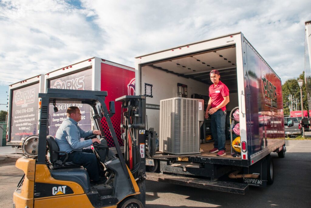 Two technicians loading an outdoor HVAC unit onto a company truck with a forklift for Parks Heating Cooling Plumbing & Electrical in Indian Trail, NC
