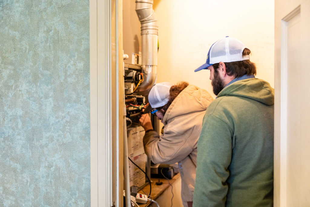 Two HVAC technicians performing repairs on an indoor furnace or air handler unit for Comfort Engineering Co Inc in Tupelo, MS