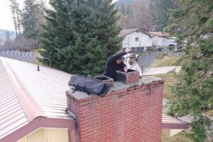 Two Chimney Wizards LLC technicians on a roof, actively working on a chimney flue during a sweep in Orofino, ID.