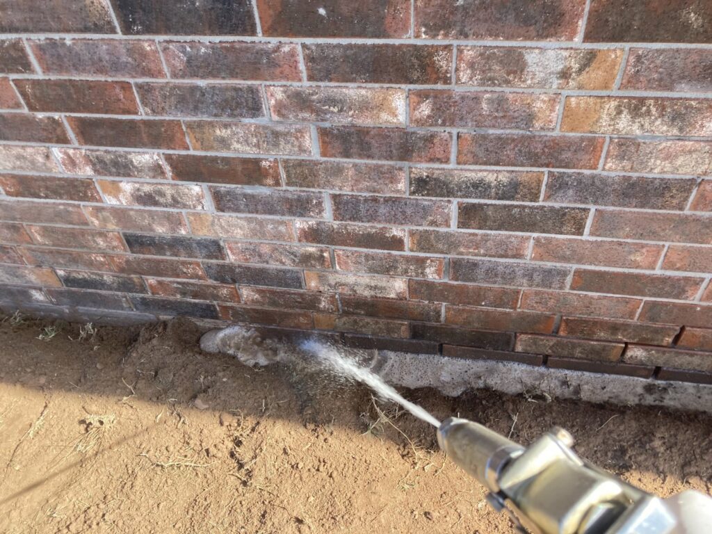Close-up of a technician spraying liquid for termite treatment into the ground next to a brick foundation by Roach's Termite Service Inc. in Oklahoma City, OK