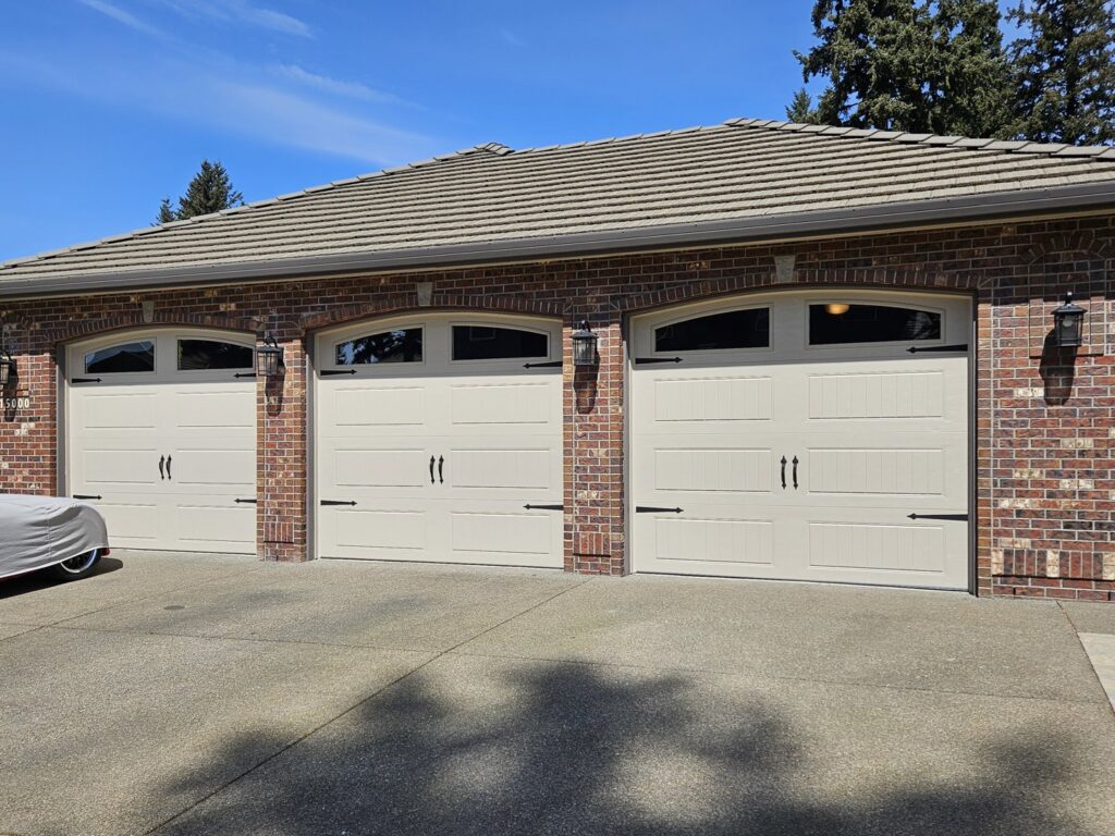 Three light-colored carriage-style garage doors with decorative hardware on a brick house by Babcock Garage Doors in Vancouver, WA