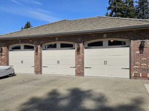 Three light-colored carriage-style garage doors with decorative hardware on a brick house by Babcock Garage Doors in Vancouver, WA