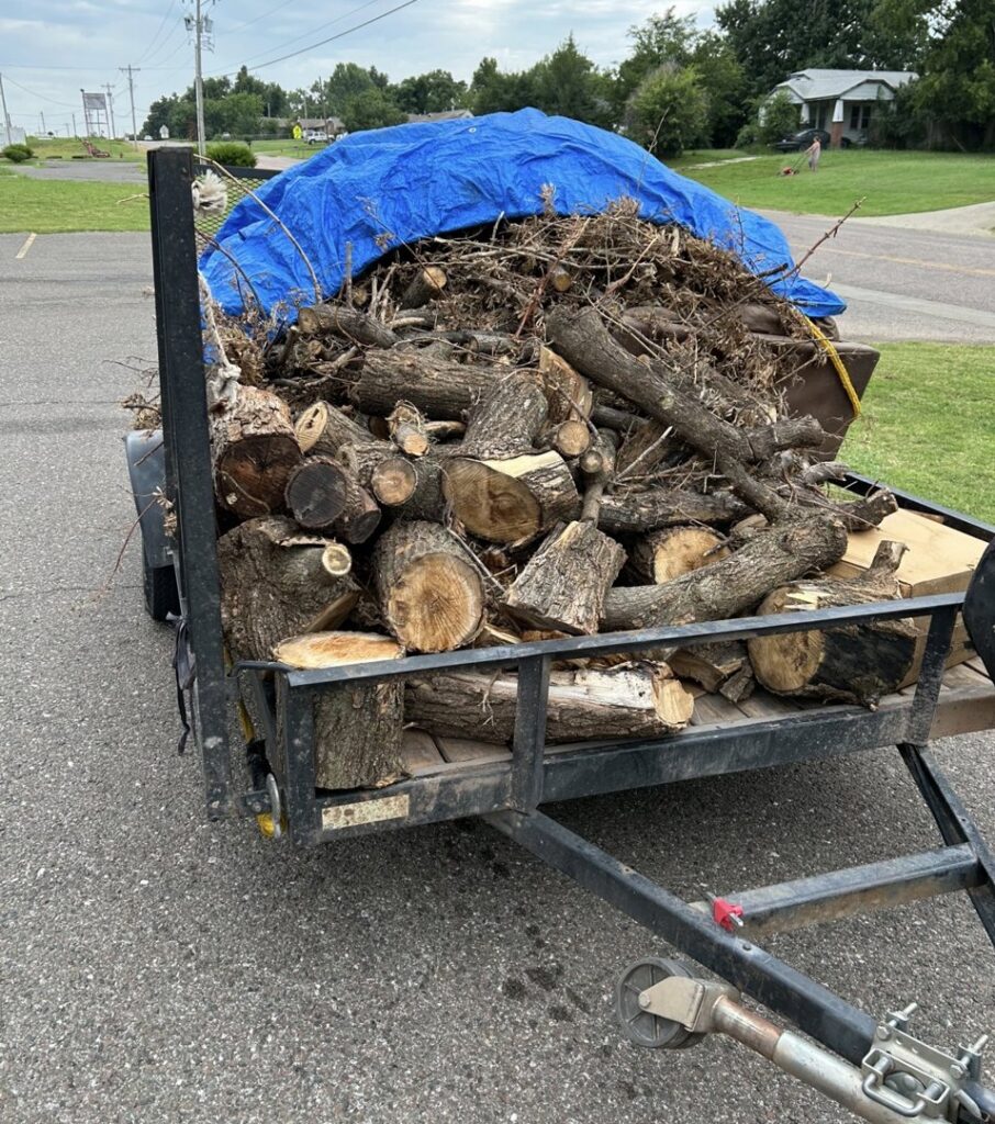 A trailer loaded with tree debris and wood logs, covered with a blue tarp, for removal by Lawns Plus More LLC in Edmond, OK.