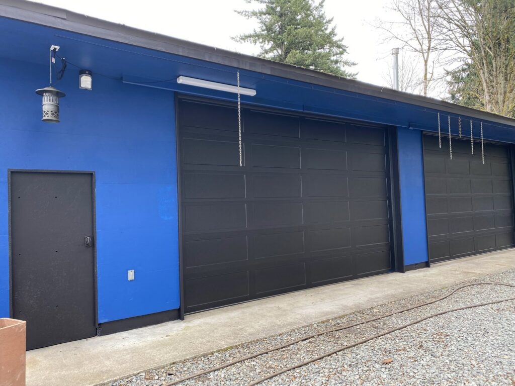 Two black panel garage doors installed on a blue commercial or industrial building by Independent Garage Doors LLC in Tacoma, WA.
