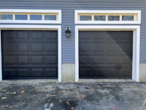 Two black garage doors with transom windows installed on a blue-sided house by PM Garage Door in Hudson, NH.