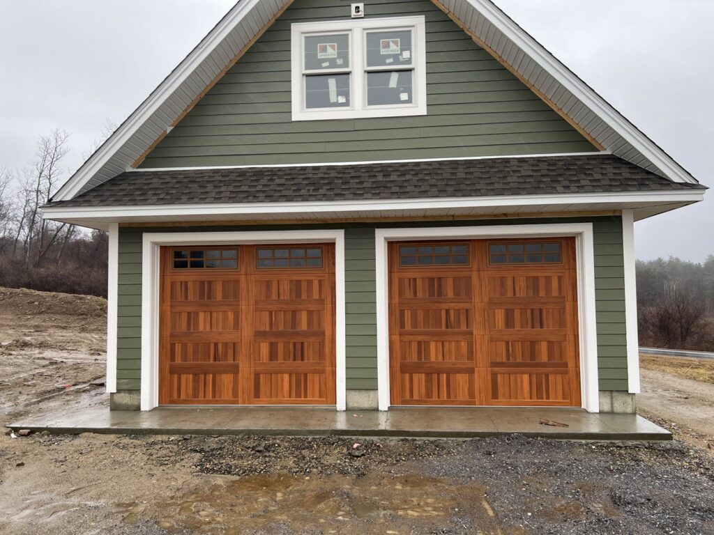 Two brown wood-grain garage doors with windows installed on a green-sided garage by PM Garage Door in Hudson, NH.